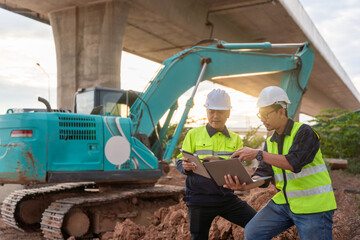 Asian two construction engineers review plans on a laptop with a large excavator and a concrete overpass in the background.