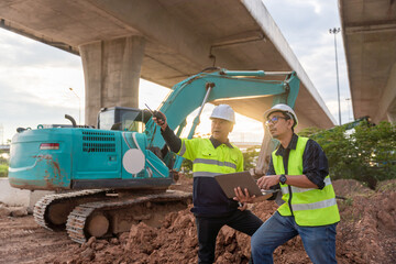 Asian two construction engineers use a radio and laptop to direct an excavator under a towering highway overpass at sunset.