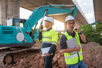Asian Two construction engineers stand on a dirt pile, using radios and tablets to supervise work near an excavator at sunset.