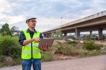 A focused Asian civil engineer works on a laptop, standing by a dusty road with a major highway overpass in the background.