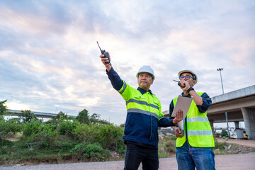 Two construction engineers use two-way radios and a tablet to communicate and point at the site under a dramatic cloudy sky.