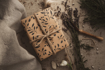 Rustic gift wrapped in botanical kraft paper and twine, styled with herbs and petals on a cozy fabric backdrop. Warm artisanal holiday packaging flat lay.
