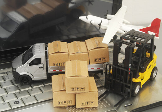 Truck model, forklift and airplane with many carton boxes on laptop computer close up. Cargo and online business concept.