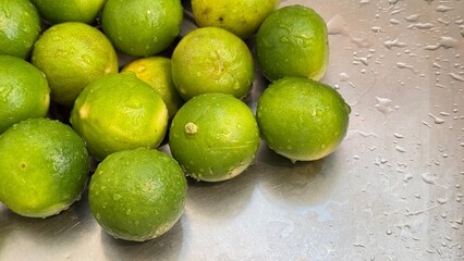 Fresh green limes gathered on a stainless-steel surface, with water droplets on both the limes and the surrounding area, giving a clean and refreshing appearance