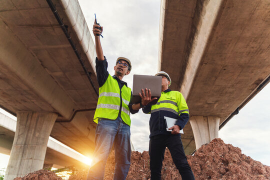 Construction workers using technology on site under highway overpass with sunlight casting shadows in urban environment during project planning phase