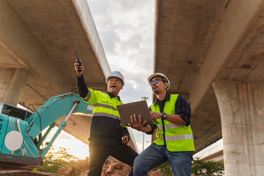 Two construction workers discussing project details under a highway bridge with heavy machinery in the background during sunset