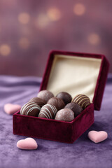 Assorted chocolate truffles in red velvet box with pink heart shaped candies on purple fabric background, creating romantic and elegant gift presentation