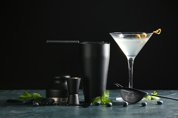 Bartender utensils and glass with cocktail and ingredients on table near black wall, closeup