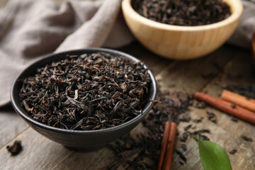 Bowls with dry black tea leaves and cinnamon on wooden background, closeup