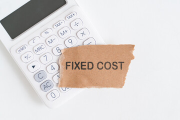 FIXED COST text on grunge uneven torn recycled pater and blurred calculator above white desk, top view