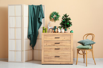 Interior of living room with folding screen, chair, Christmas tree, gift boxes and candles on commode near beige wall