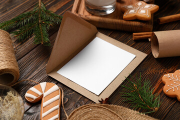 Blank greeting card with envelope, Christmas decorations, gingerbread cookies and packing materials on brown wooden background