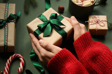 Female hands with Christmas decorations, cup of cacao, candy cane and gift boxes on green background