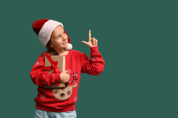 Obraz premium Cute little girl in Santa hat with gift book pointing at something on green background