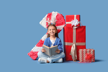 Naklejka premium Cute little girl with book and Christmas gifts sitting on blue background