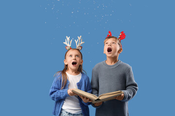 Naklejka premium Surprised little children in Christmas reindeer horns with book and snow on blue background