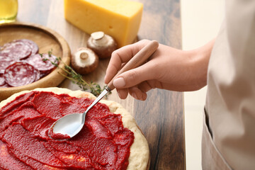 Woman with ingredients spreading tasty tomato sauce on raw pizza dough in kitchen, closeup