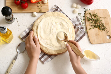 Woman with ingredients spreading tasty white sauce on raw pizza dough in kitchen, top view