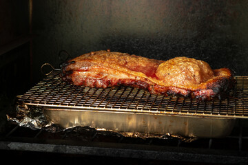 Thick, savory pork belly or crispy roast cooking on a wire rack over a drip pan inside a dark oven or smoker, with a skewer holding the cut. Focus on the golden, crackling skin.