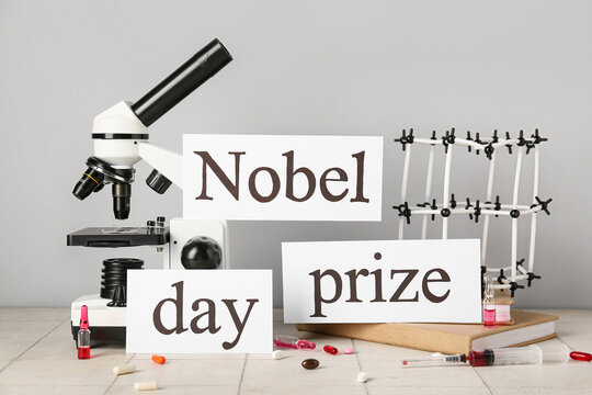 Paper with text NOBEL PRIZE DAY, molecular model, microscope, books and pills on tile against grey background