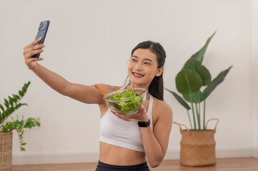 Healthy young Asian woman in sportswear eating fresh salad after workout. She finished exercising in her living room at home.