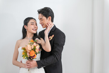 Romantic Asian bride and groom posing together indoors. The groom in a black suit gently embraces the bride in a white wedding dress and holding flowers