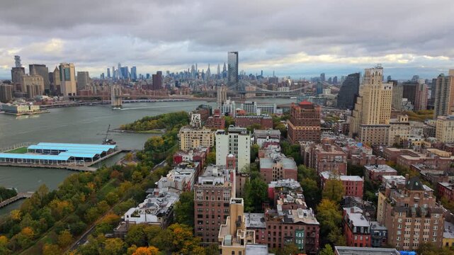 Aerial drone footage of New York City, Brooklyn, New York showing Brooklyn Bridge, river surface, green waterfront areas and distant skyline towers under cloudy daylight. United States