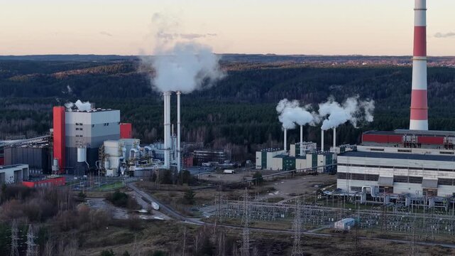 Drone shot with a slight panning movement showing a cogeneration power plant in Vilnius, white smoke rising from multiple chimneys, large structures and the forest in the sunrise light.