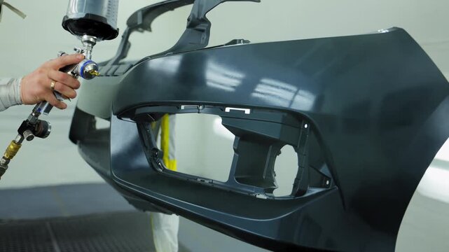 Auto technician spray paint or lacquer on car bumper inside clean, ventilated paint booth. Wearing protective coveralls and a respirator using a high precision spray gun.