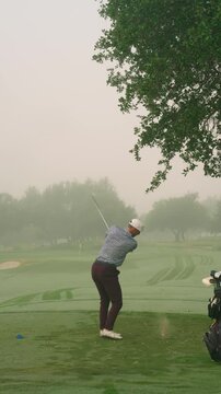 Vertical Behind-angle shot of male golfer hitting iron off tee on foggy morning, pin visible as he finishes with a smooth club twirl beside his golf bag