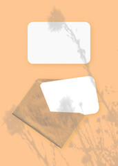 The shadow of wildflowers falls on an envelope with two sheets of textured white paper on a orange background. Mockup vertical orientation.