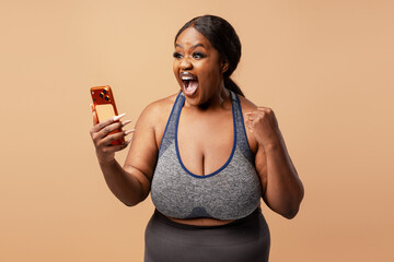 Happy African American woman holding mobile phone, using mobile app win money on beige background
