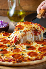 Delicious Pepperoni Pizza Slice Being Lifted with a Spatula, Showcasing Melted Cheese and Toppings on a Wooden Board with Olive Oil and Red Cabbage in Background