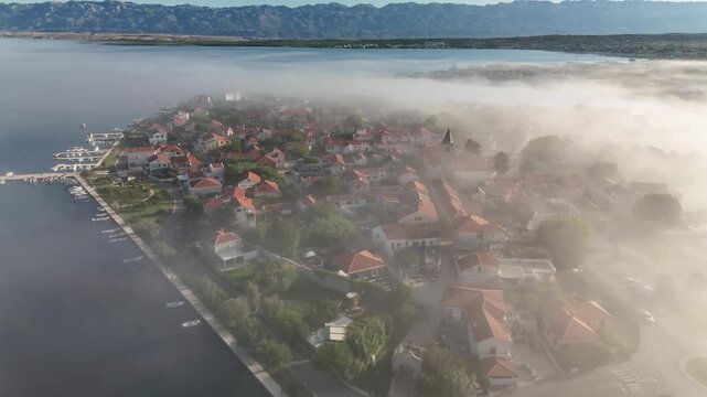 A peaceful aerial sunrise over Nin, Croatia. Misty morning light reveals the old island town, stone walls, bridges, salt fields and the Velebit range as the drone gently ascends.