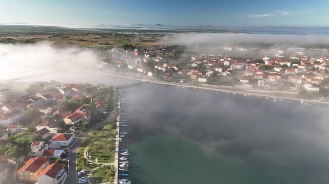 The drone turns and glides above the edge of the island, revealing the narrow sea strait that separates Nin, Croatia from the mainland. Morning mist drifts across the water as the sun breaks through
