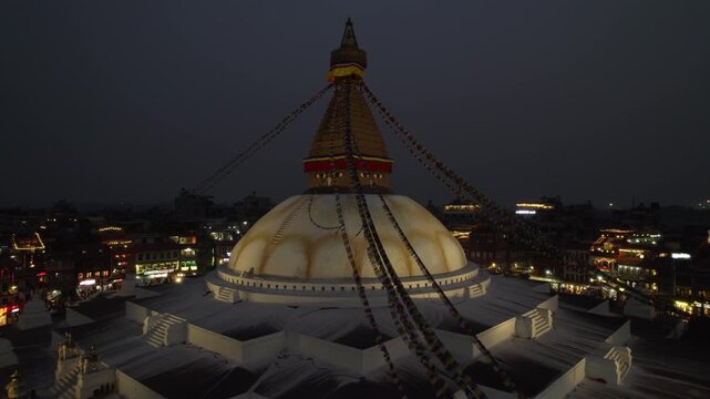 A calm night drone view of Boudhanath Stupa, with boudha and nath noted separately, glowing above Kathmandu. The sacred Buddhist landmark stands radiant in the quiet evening cityscape.