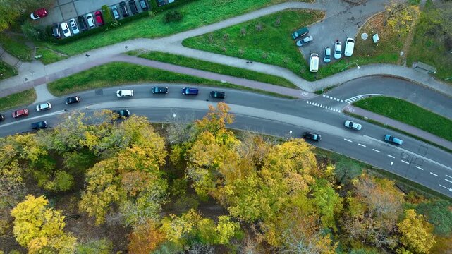 Overhead drone shot of cars driving on a curved road in Gdansk next to a park and parking area during the autumn season.
