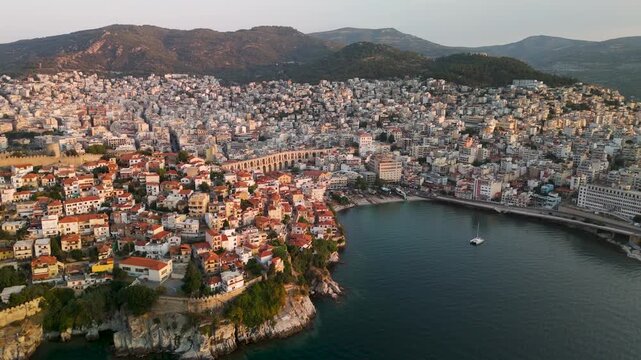 Sweeping wide aerial drone approach over the Thracian Sea towards the sprawling coastal city of Kavala, Eastern Macedonia, Greece, illuminated by the rising sun.