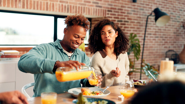 Happy couple, family and juice with food for dining, meal or gathering together at dinner table. Man, woman or pouring with orange drink or smile for vitamin C, thanksgiving or serving in house - Powered by Adobe