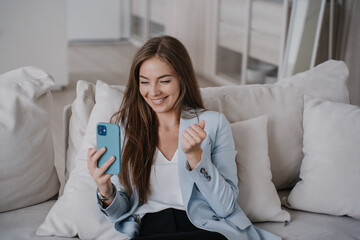 A woman relaxes on a sofa, smiling at her phone, appearing thoughtful and engaged in a stylish and airy living space.