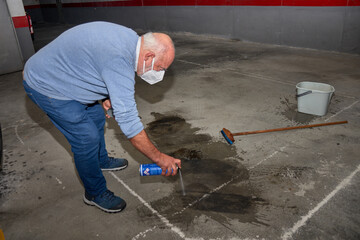 Elderly man cleaning an oil stain in a garage with spray in an editorial scene about urban maintenance.