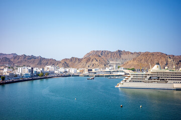 A stunning panoramic view of Muttrah Harbor in Muscat, Oman, featuring turquoise blue water, white coastal buildings, and dramatic desert mountains rising behind the city