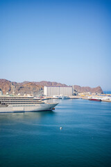 A luxury cruise ship docked at the Port of Muscat, Oman, surrounded by calm blue water and framed by rugged desert mountains. The scene includes port buildings, industrial silos, smaller vessels