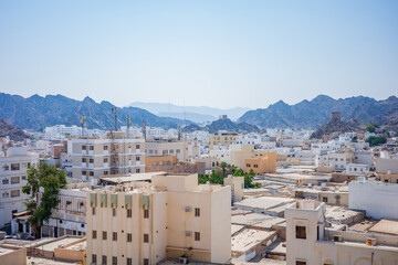 A panoramic view of Old Muscat, Oman, showcasing beige buildings, traditional Middle Eastern architecture, and rugged mountain ranges in the background. The soft blue sky and layered mountains