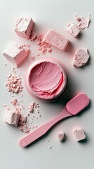 A close-up overhead view of a pink cosmetic cream in an open jar, surrounded by scattered pink powder and chunks of product, with a pink spatula lying nearby on
