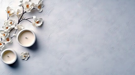 Two unlit white candles and a branch of white flowers are arranged on a textured white surface, creating a peaceful and minimalist composition.