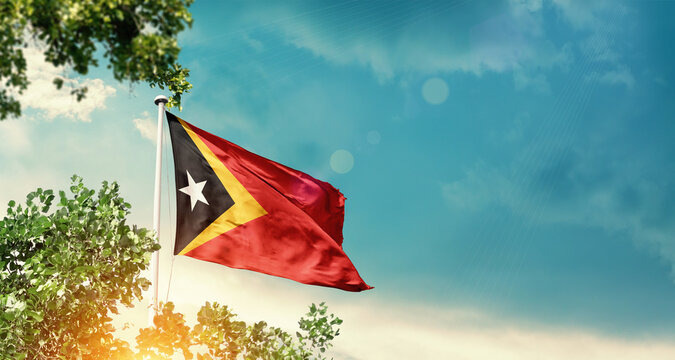 East Timor national flag waving in the sky behind the tree. The flag blowing with beautiful sunlight. East Timor national flag for independence day. - Powered by Adobe