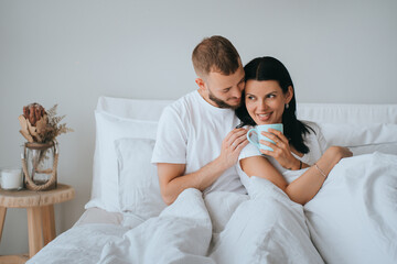 A cozy morning in bed with a couple sharing coffee under a white duvet, radiating warmth and affection in a minimalist setting.