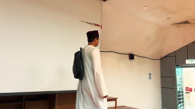 A focused Muslim student sits at a desk in a classroom, reading, thinking, and writing notes. The scene captures concentration, learning, and academic dedication in a bright educational environment. 