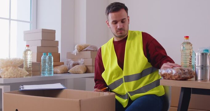Man volunteer packs food products in cardboard box, writing something down and holding clipboard, prepares humanitarian aid for poor people, works in charitable foundation or charity centre